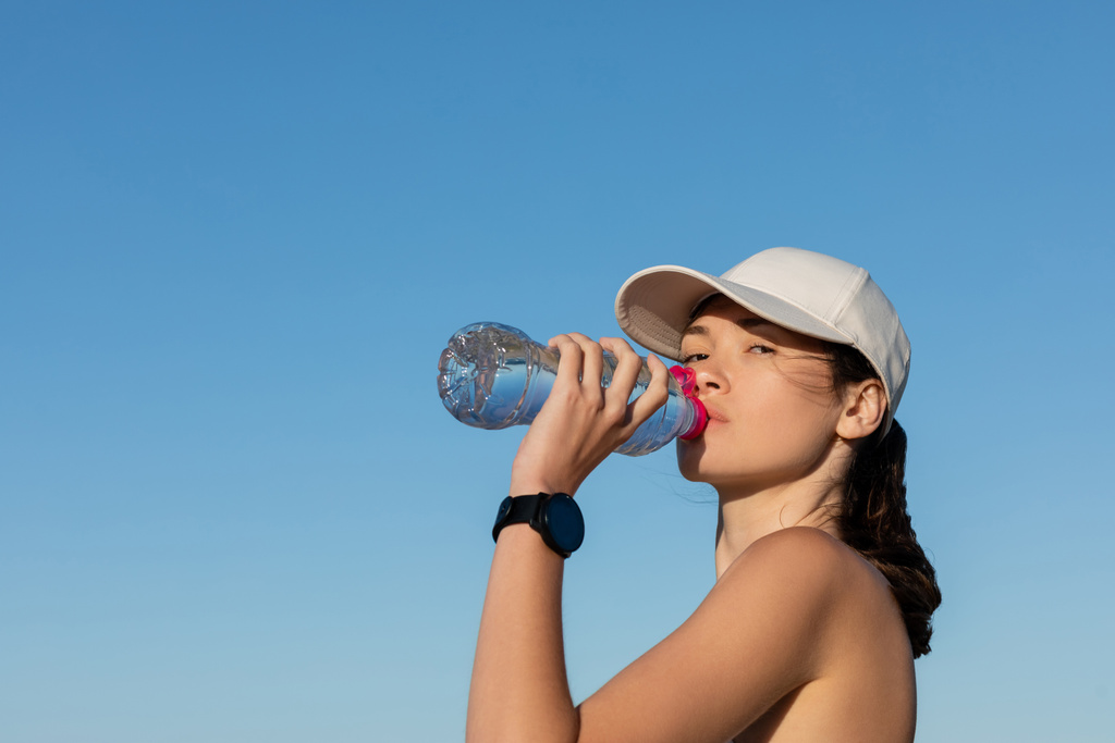 Sportliche Frau mit Baseballmütze trinkt erfrischendes Wasser gegen blauen Himmel - Foto, Bild
