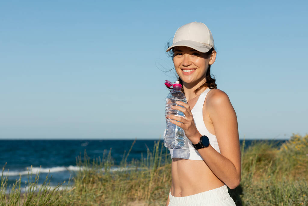 Beyzbol şapkalı, mutlu ve sportif bir kadın elinde okyanus kenarında suyla şişe tutuyor. - Fotoğraf, Görsel