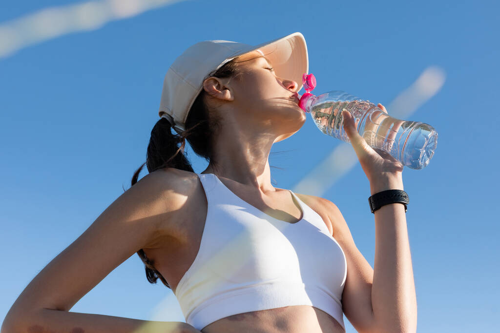vue à angle bas de la femme sportive en casquette de baseball et soutien-gorge de sport boire de l'eau contre le ciel bleu - Photo, image