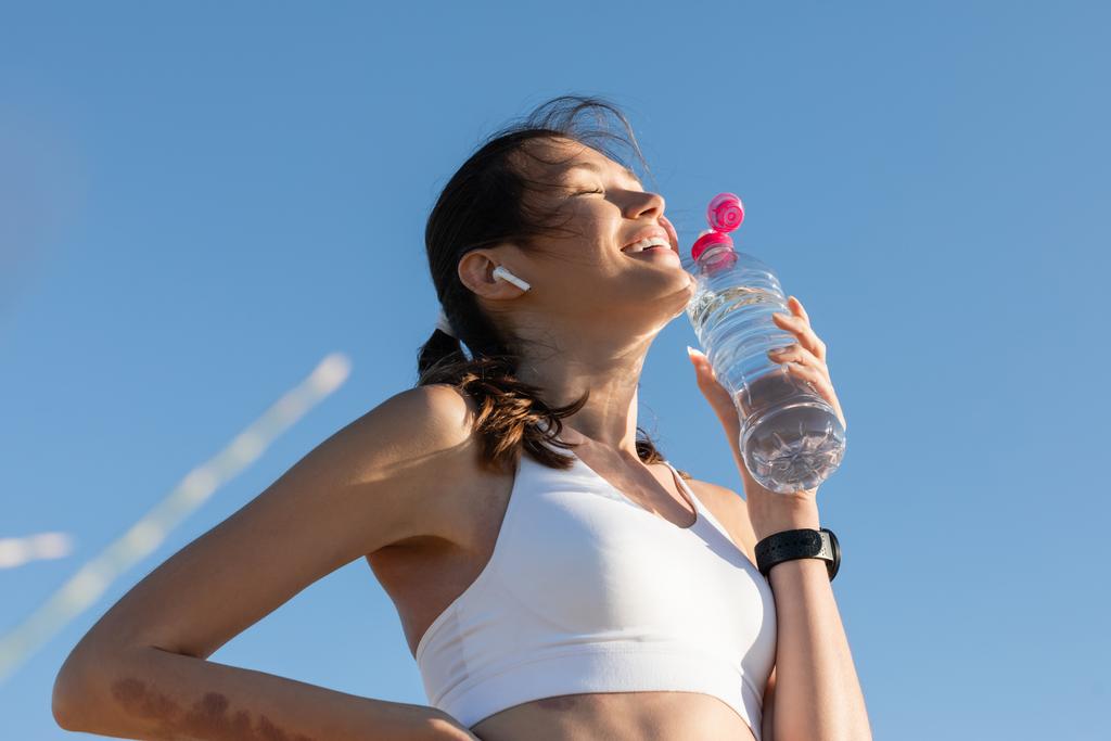 Tiefansicht einer fröhlichen Frau im drahtlosen Kopfhörer, die eine Flasche mit Wasser gegen den blauen Himmel hält - Foto, Bild