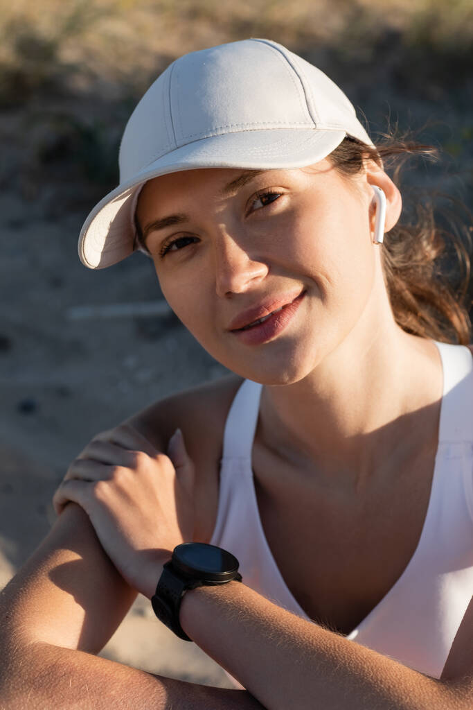 portret van gelukkige sportvrouw in baseball cap luisteren muziek in draadloze oortelefoon op het strand - Foto, afbeelding