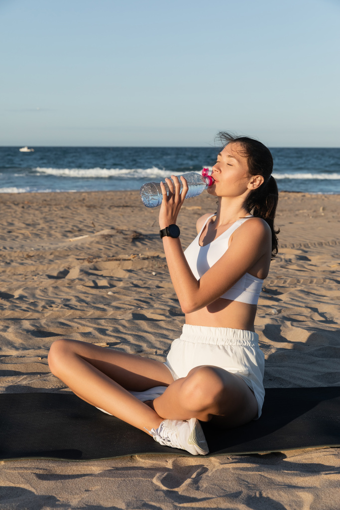 молода жінка тримає пляшку з водою, сидячи на фітнес-маті біля моря
 - Фото, зображення