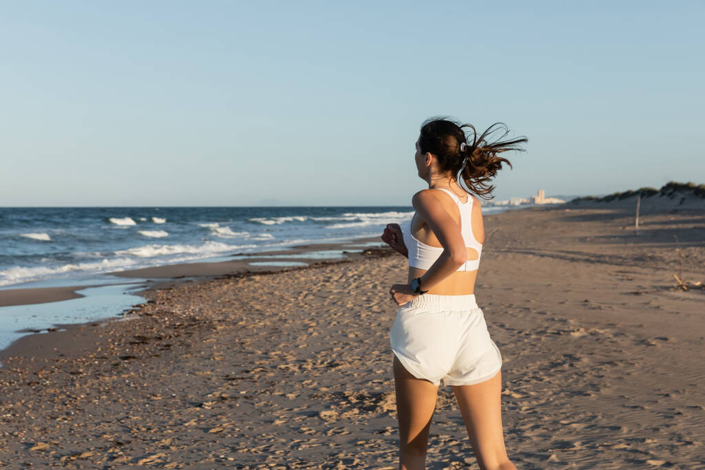 молодая брюнетка в белом спортивном костюме бегает на песчаном пляже летом - Фото, изображение
