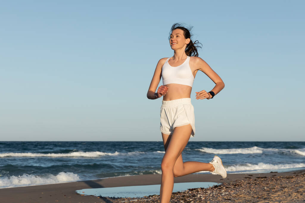 Lächelnde junge Frau in Sportkleidung joggt im Sommer am Meer - Foto, Bild