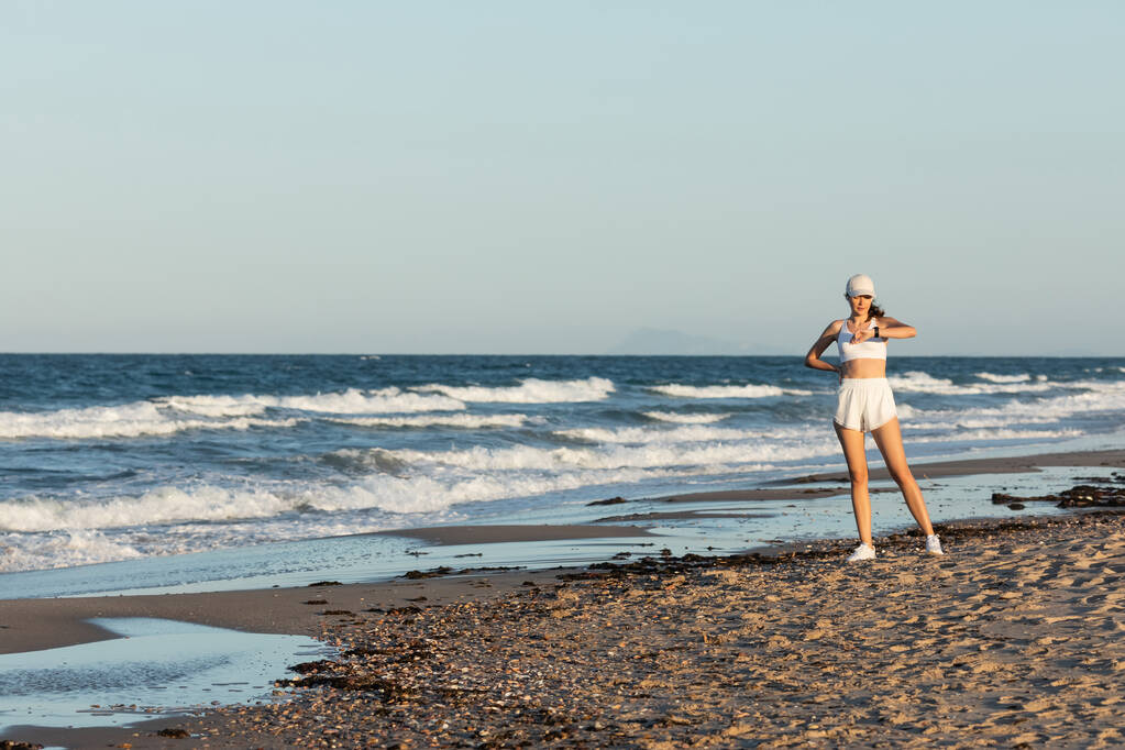mladá žena v baseballové čepici při pohledu na fitness tracker v blízkosti moře na pláži - Fotografie, Obrázek