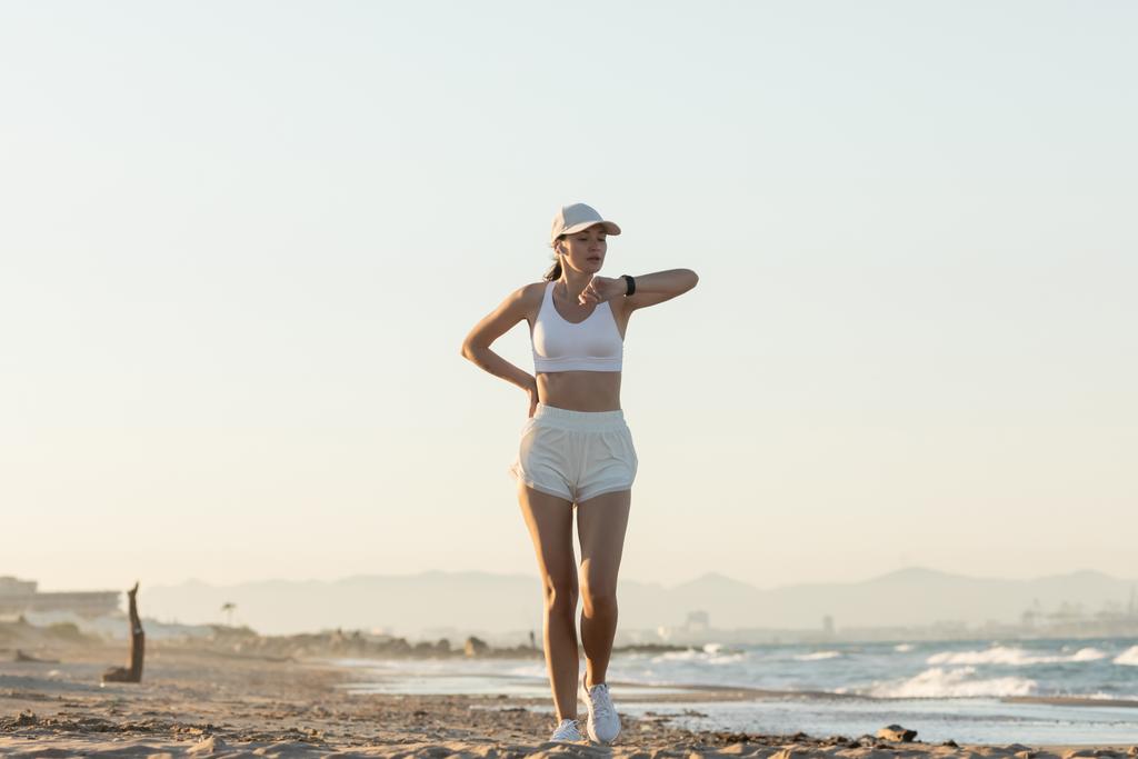 teljes hossza fiatal nő baseball sapkában nézi fitness tracker közel tenger  - Fotó, kép