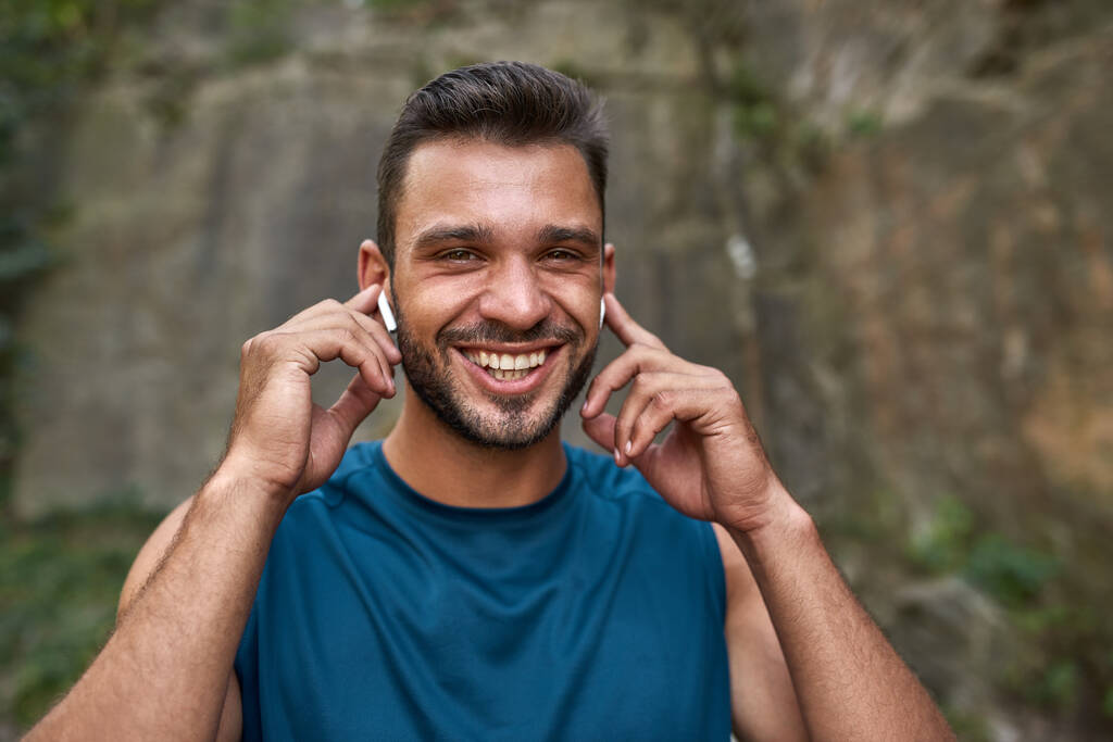 Портрет улыбающегося молодого кавказского спортсмена, выбирающего музыку в беспроводных наушниках на открытом воздухе. Красивый бородатый мужчина в спортивной одежде смотрит в камеру. Концепция современного здорового образа жизни - Фото, изображение