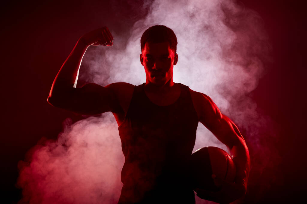 Strona koszykarza Silhouette oświetlona czerwonym kolorem trzymając piłkę z dymem w tle. Poważny Afrykański Amerykanin wykazujący mięśnie - Zdjęcie, obraz