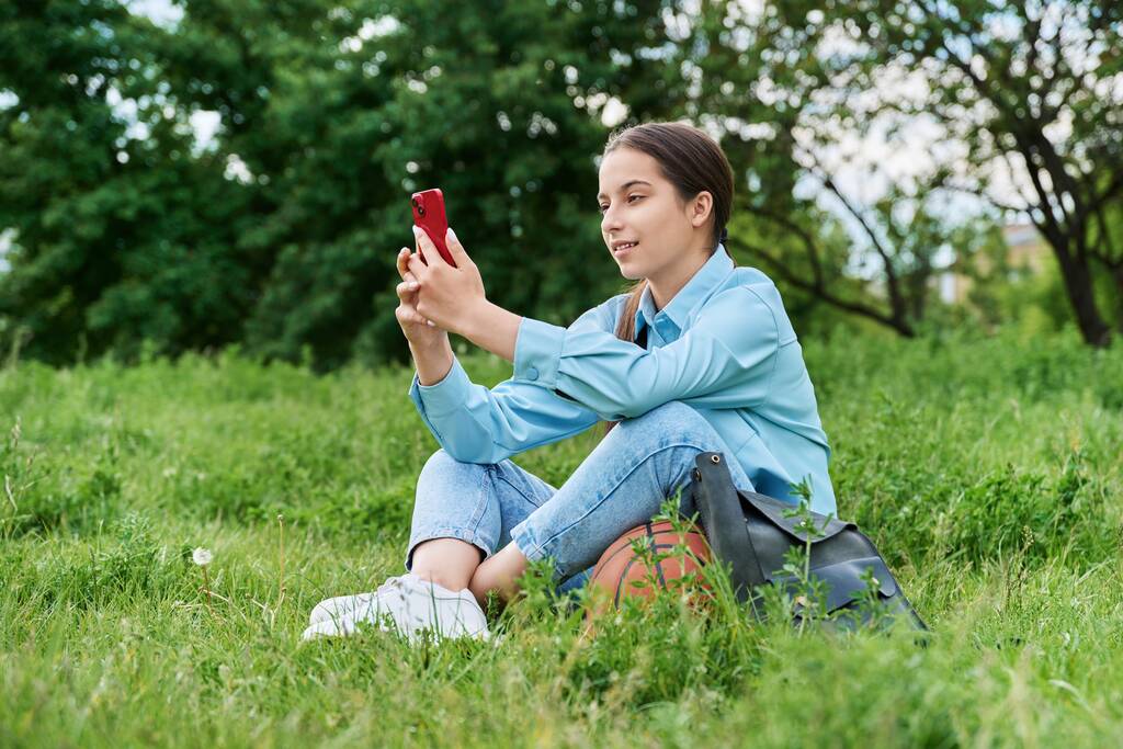 Adolescente estudiante que descansa sentada en la hierba usando un teléfono inteligente. Atractiva chica adolescente en el césped con pelota de baloncesto, mochila. Escuela secundaria, tecnología, estilo de vida activo y saludable, concepto de adolescencia - Foto, imagen