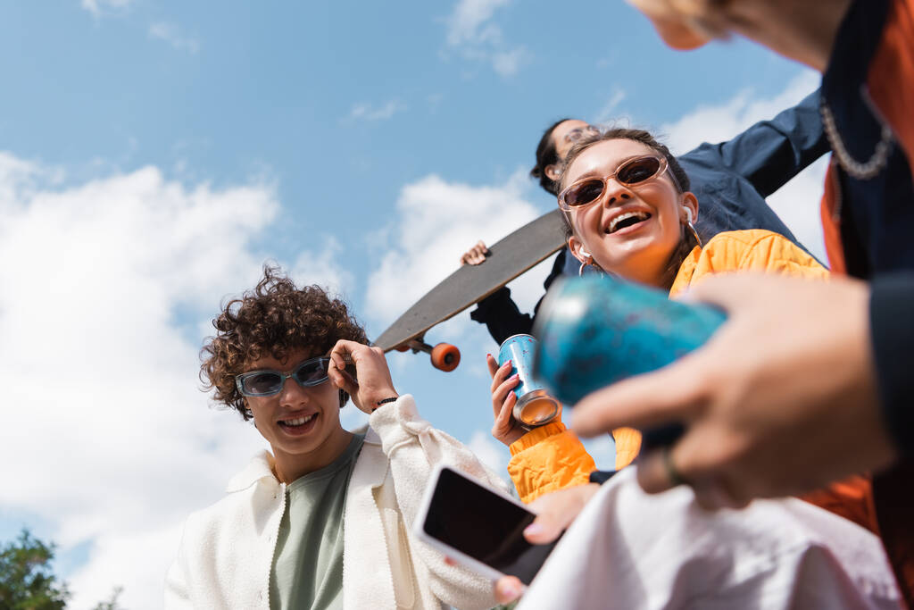 vista de ángulo bajo de alegres amigos multiétnicos con lata de refrescos y teléfono inteligente al aire libre - Foto, Imagen