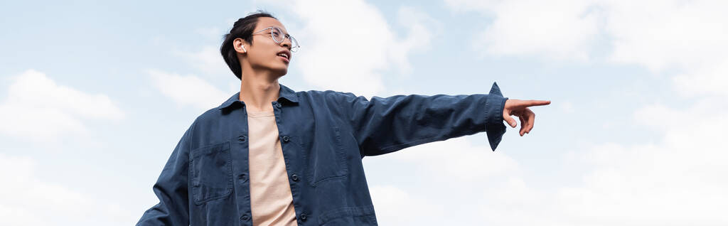 low angle view of asian man in eyeglasses and earphone pointing with finger outdoors, banner - Photo, Image