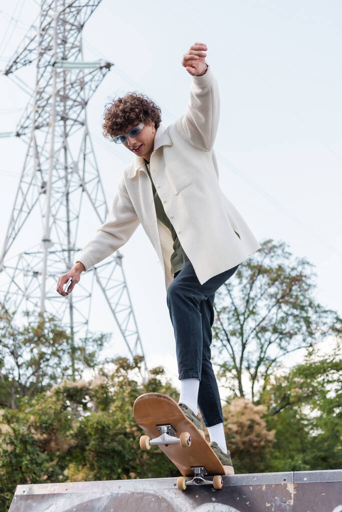 vista basso angolo di giovane uomo riccio in giacca bianca skateboard in skate park - Foto, immagini