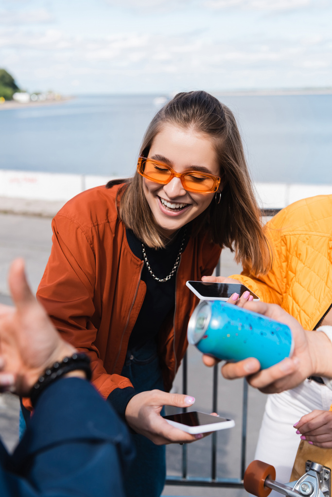 smiling woman in trendy sunglasses holding smartphone with blank screen near friends - Photo, Image