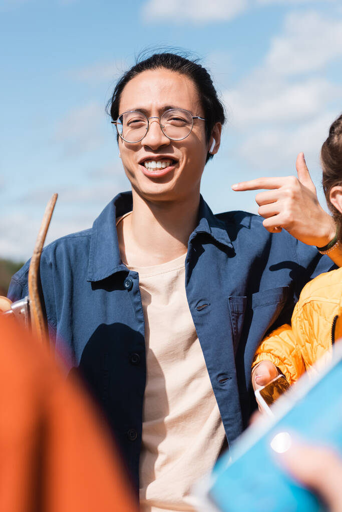 щасливий азіатський чоловік в окулярах вказує пальцем біля розмитих друзів на відкритому повітрі
 - Фото, зображення