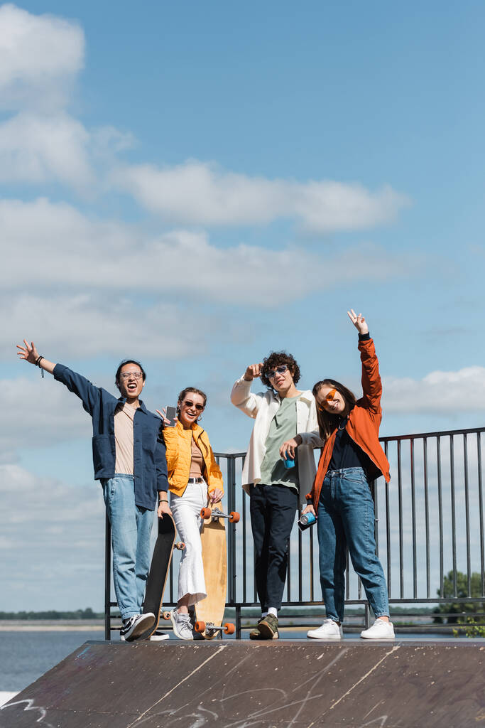 šťastný multikulturní bruslaři mávající rukama a ukazující znamení vítězství na skate rampě pod modrým oblačným nebem - Fotografie, Obrázek