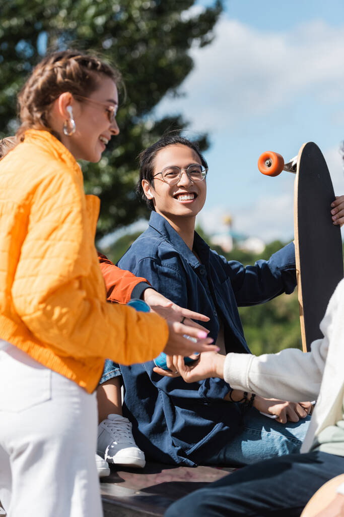 gelukkig aziatische man in bril houden longboard in de buurt van vrienden buiten - Foto, afbeelding