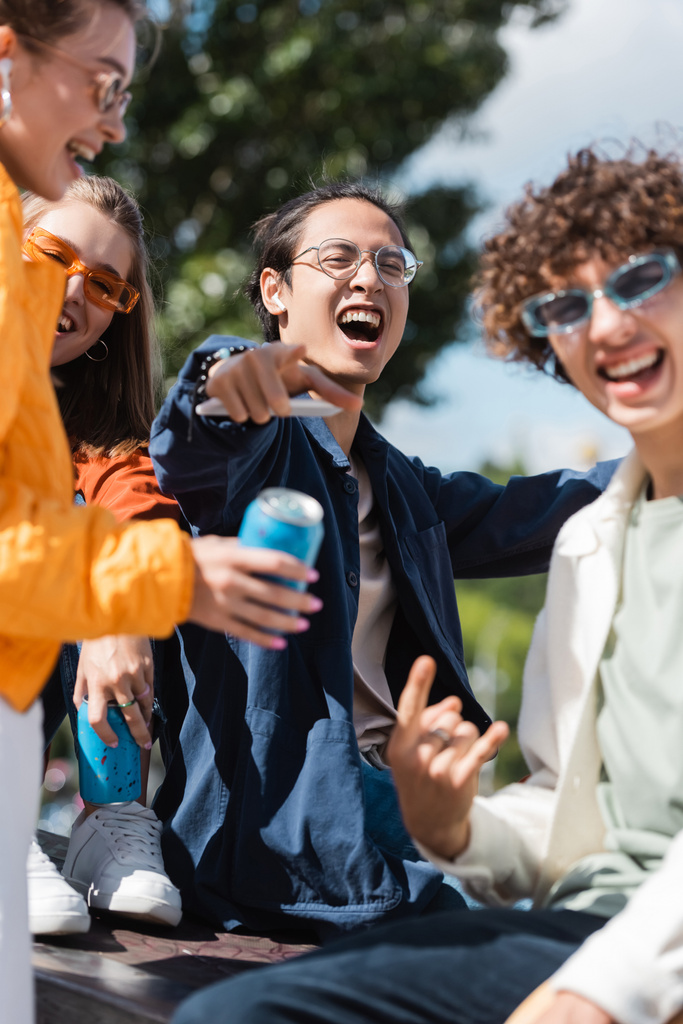 excitado asiático hombre en gafas apuntando con dedo cerca borrosa amigos al aire libre - Foto, Imagen