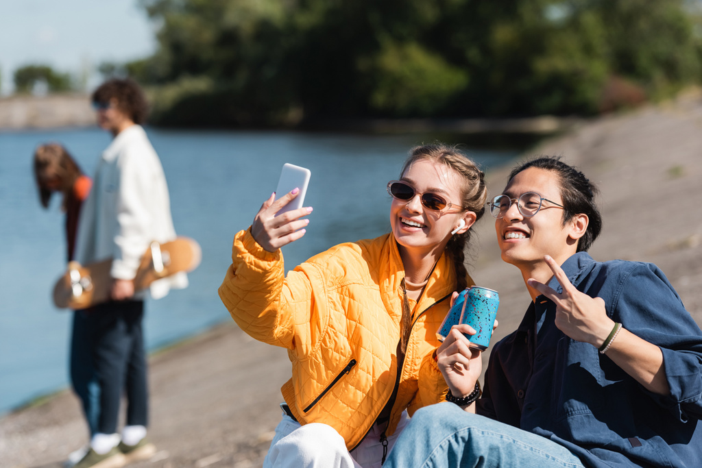 onnellinen rotujenvälinen ystäviä clinking sooda tölkit ja ottaen selfie joen rannalla  - Valokuva, kuva