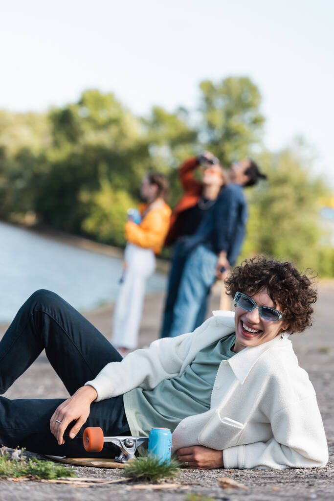 hombre rizado en gafas de sol de moda sonriendo a la cámara cerca de monopatín y amigos sobre fondo borroso - Foto, imagen