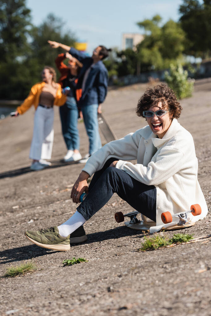 excited man in sunglasses sitting on longboard and laughing at camera outdoors - Photo, Image