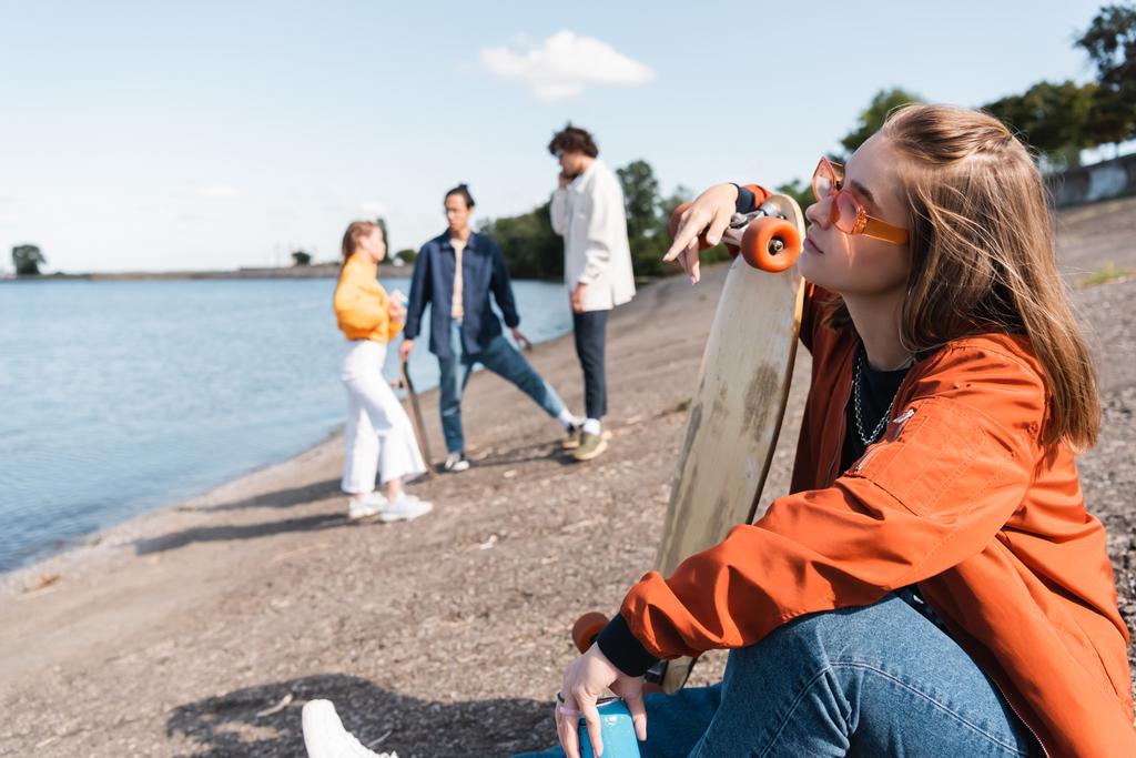 joven mujer en gafas de vista sentado en la orilla del río con penny board cerca de amigos sobre fondo borroso - Foto, imagen