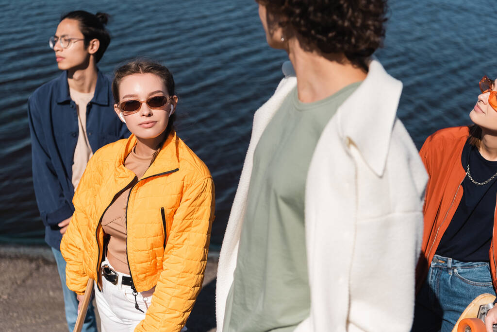 jeune femme dans des lunettes de soleil à la mode regardant la caméra près d'amis interracial  - Photo, image