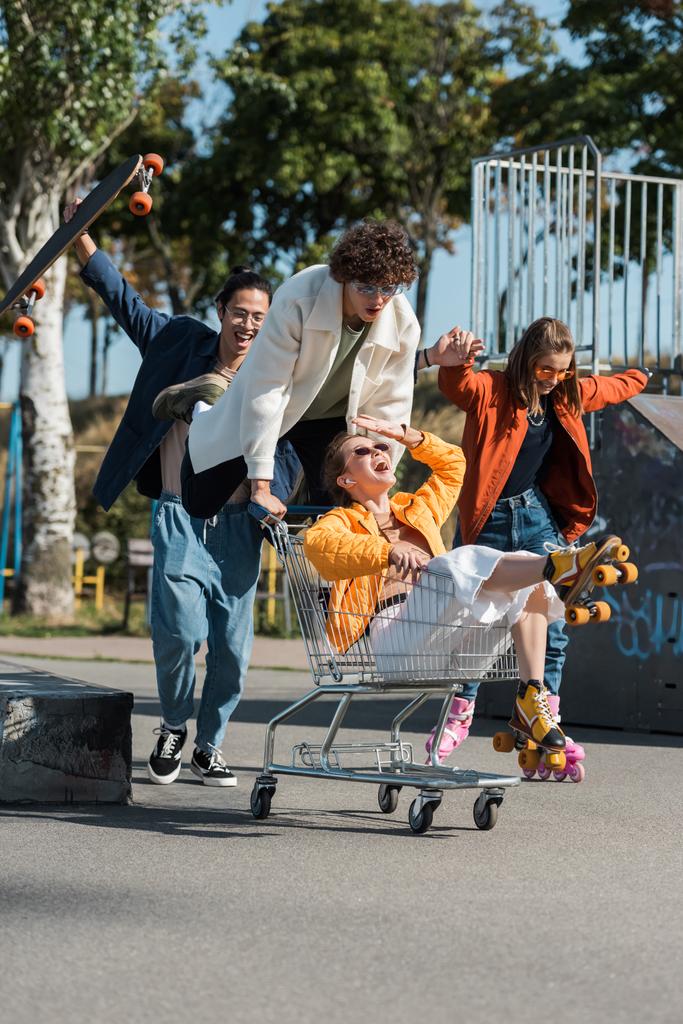 ενθουσιασμένοι πολυεθνικοί φίλοι που διασκεδάζουν με το καλάθι αγορών στο πάρκο skate - Φωτογραφία, εικόνα