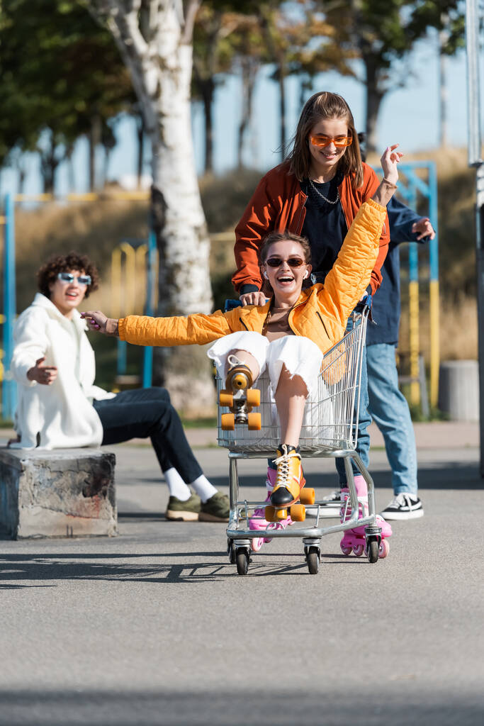 смішна жінка в роликових ковзанах катається в кошику біля щасливих багатоетнічних друзів
 - Фото, зображення