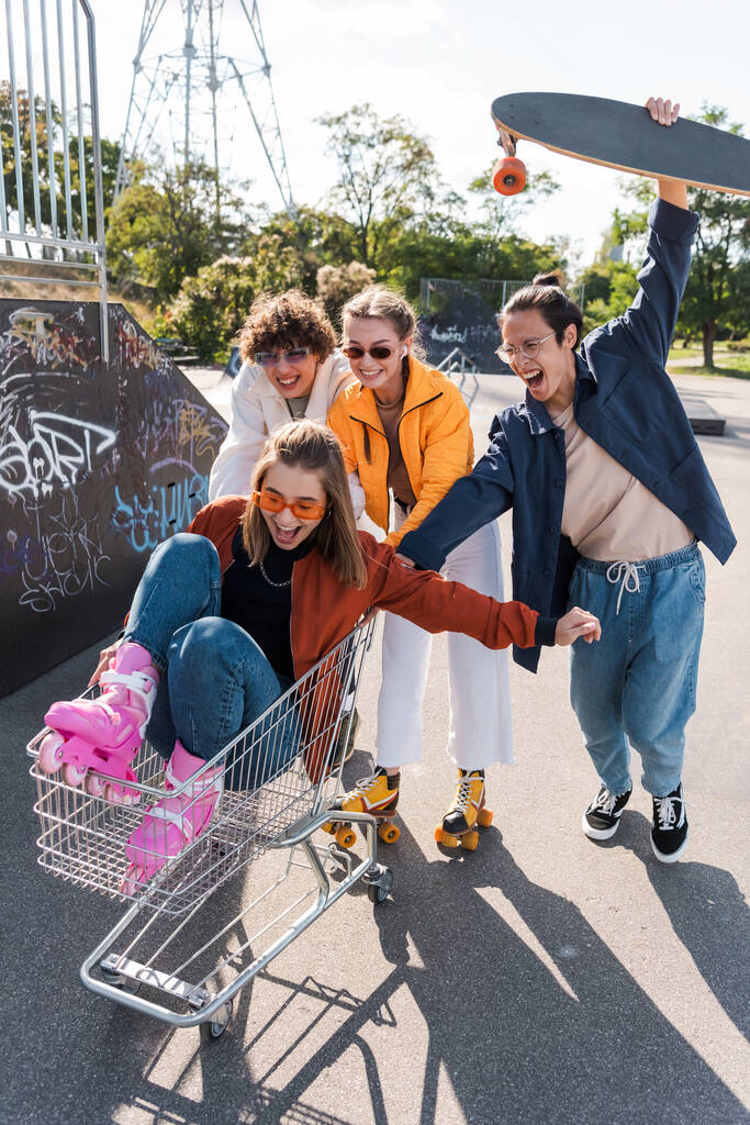 excited women shouting in shopping cart near interracial friends having fun in skate park - Photo, Image