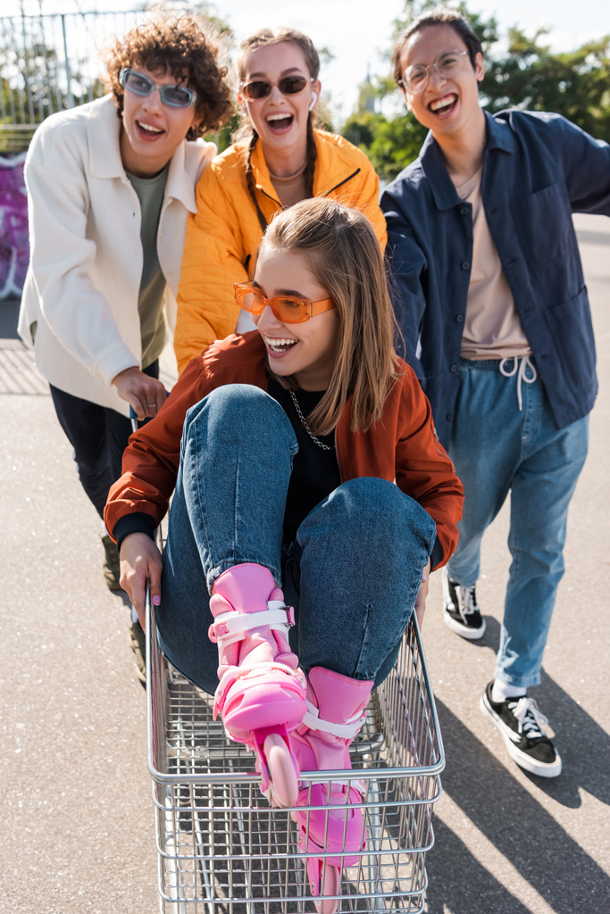 fröhliche multikulturelle Freunde bewegen Einkaufswagen mit fröhlicher Frau mit Sonnenbrille - Foto, Bild