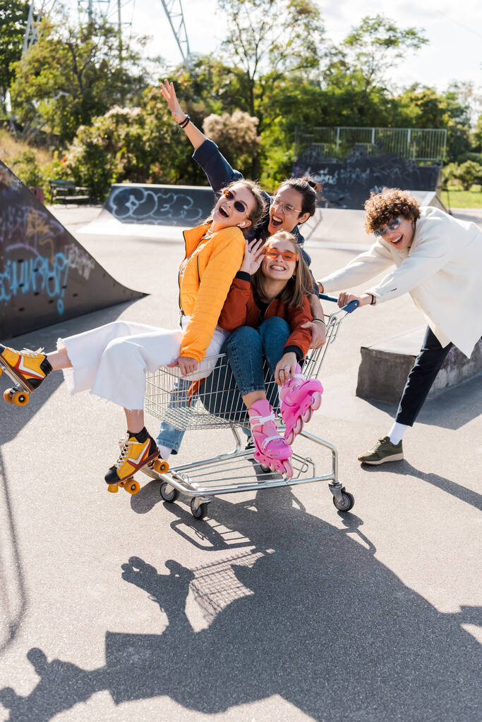 excited multiethnic friends shouting while riding in shopping cart in skate park - Photo, Image