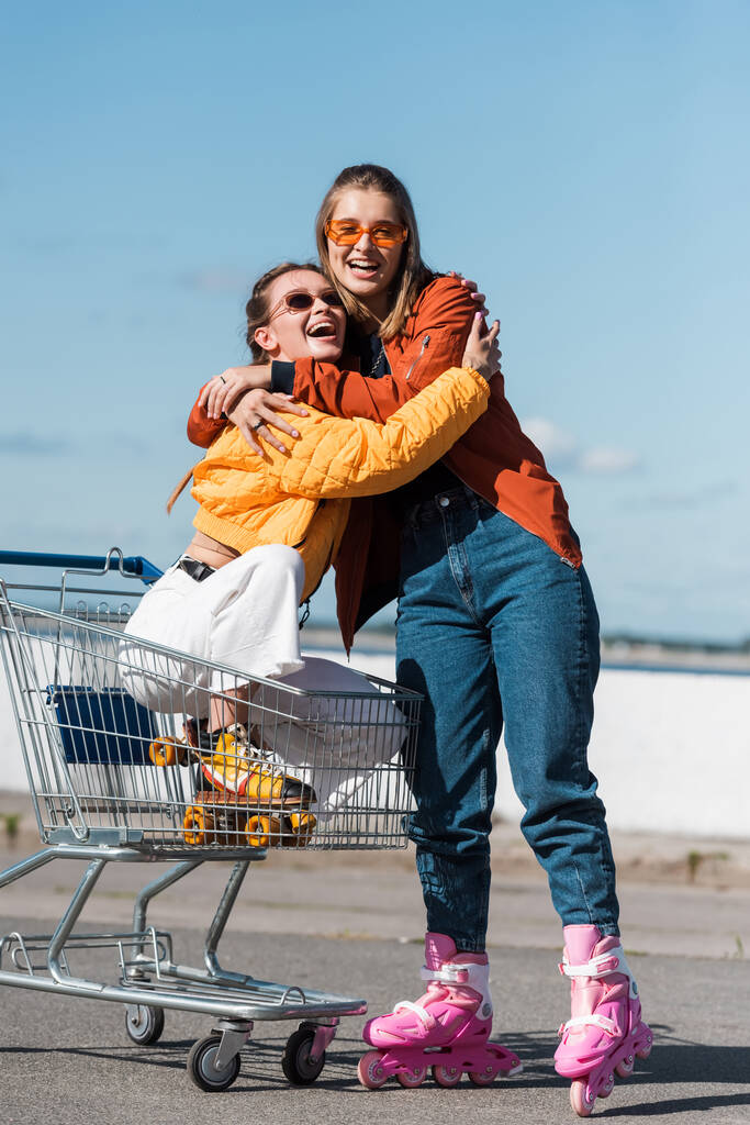 Fröhliche Frau auf Rollschuhen umarmt Freundin, die im Einkaufswagen im Freien sitzt - Foto, Bild