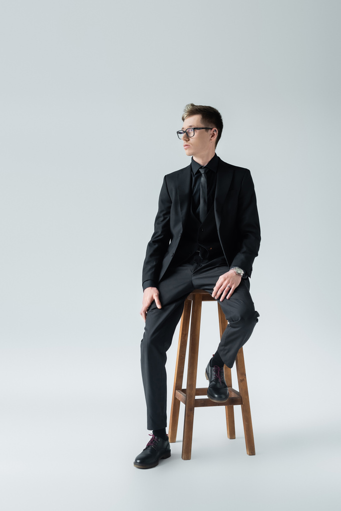 Volle Länge des eleganten Mannes mit Brille sitzt auf einem Stuhl auf grauem Hintergrund - Foto, Bild