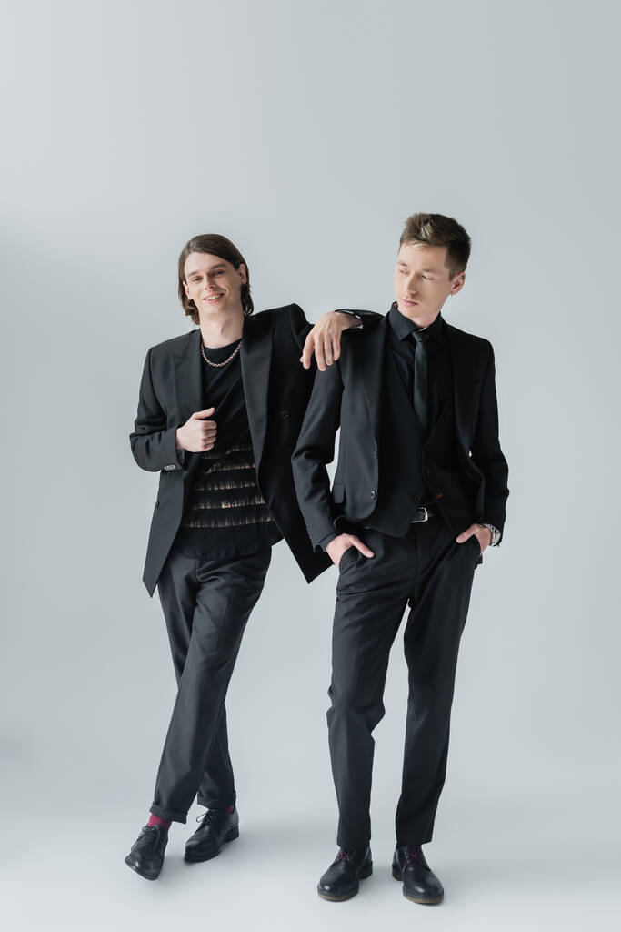 Улыбающийся гей в формальной одежде стоит рядом с парнем на сером фоне  - Фото, изображение