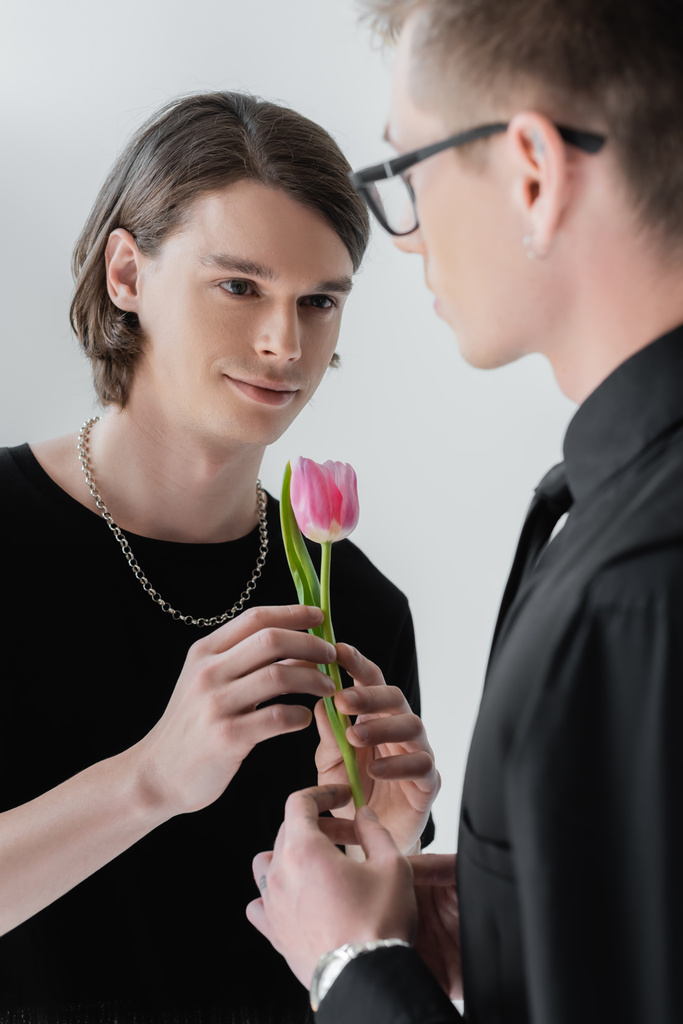 Smiling young gay man holding tulip with blurred boyfriend isolated on grey  - Photo, Image