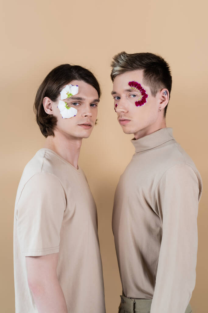 Fiatal homoszexuális pár szirmokkal az arcán, akik a bézsen izolált kamerát nézik  - Fotó, kép