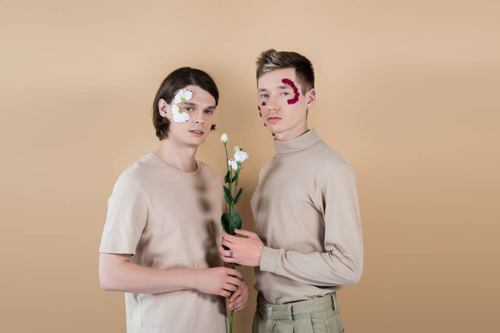Молода пара з квітковими пелюстками на обличчях тримає еустому на бежевому
  - Фото, зображення