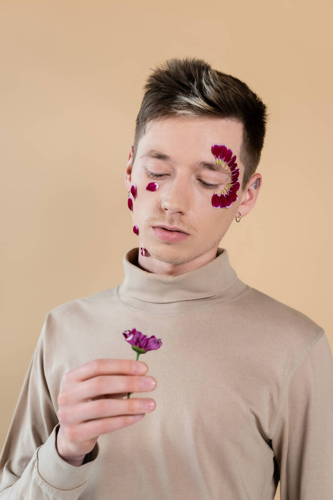 Mann mit Blütenblättern im Gesicht hält Chrysanthemenblüte isoliert auf beige  - Foto, Bild