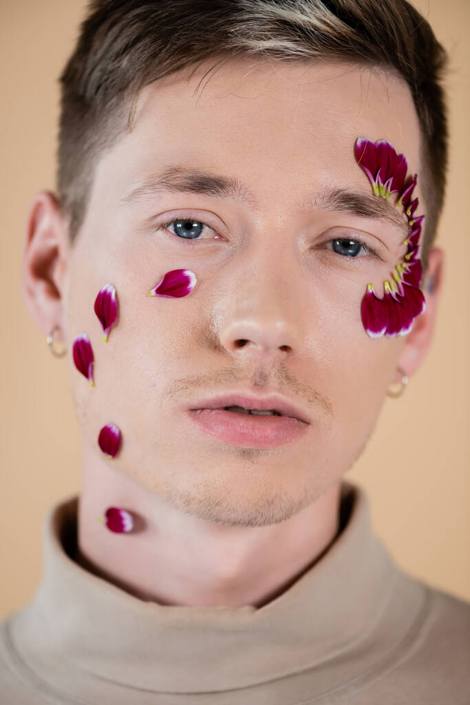 Porträt eines jungen Mannes mit Blütenblättern im Gesicht, der isoliert auf Beige in die Kamera blickt  - Foto, Bild