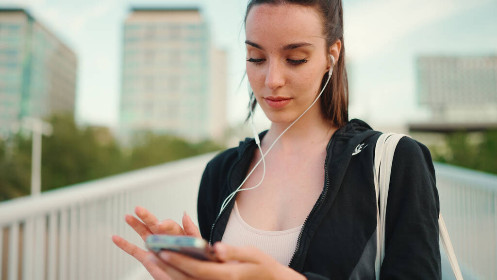 Nuori nainen pisamia ja pitkä poninhäntä yllään musta huppari ja beige urheilu top käyttää matkapuhelinta ja kuuntelee musiikkia seistessään sillalla moderni kaupungin tausta - Valokuva, kuva