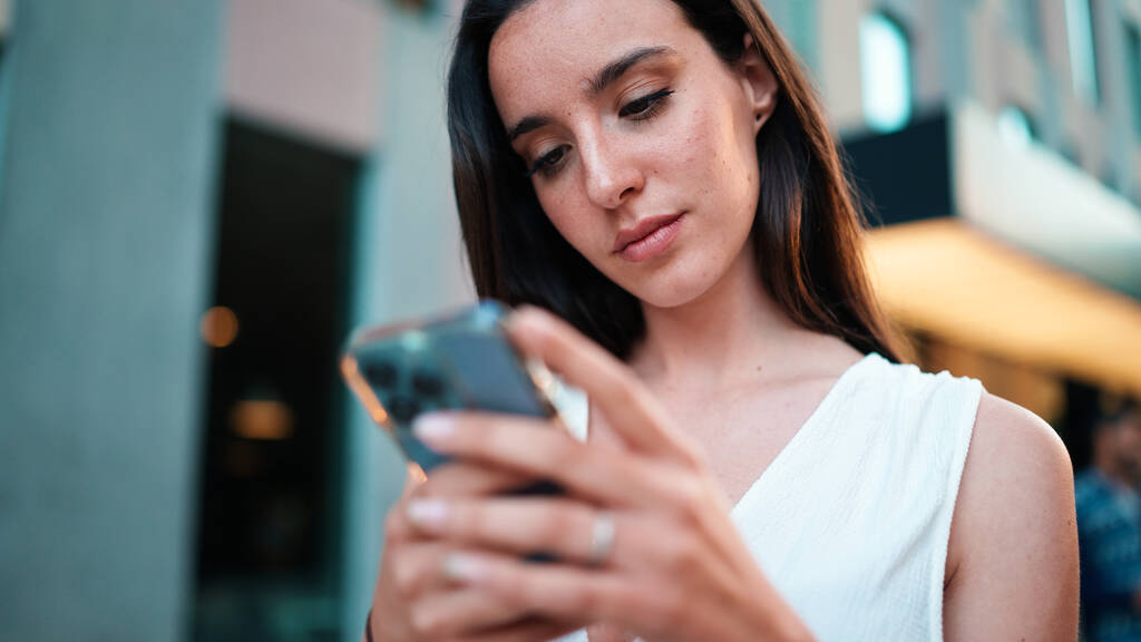 Όμορφη γυναίκα με φακίδες και σκούρα χαλαρά μαλλιά φορώντας λευκό μπλουζάκι περπατά στο δρόμο με smartphone στα χέρια της. Κορίτσι χρησιμοποιεί το κινητό τηλέφωνο στο σύγχρονο φόντο της πόλης - Φωτογραφία, εικόνα