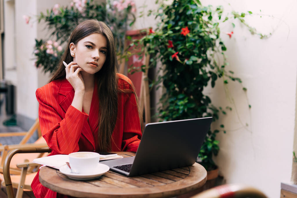 Nuori nainen freelancer kaunis punainen puku kynä kädessä istuu pöydässä kannettava tietokone ja kuppi kuumaa juomaa työskennellessään - Valokuva, kuva