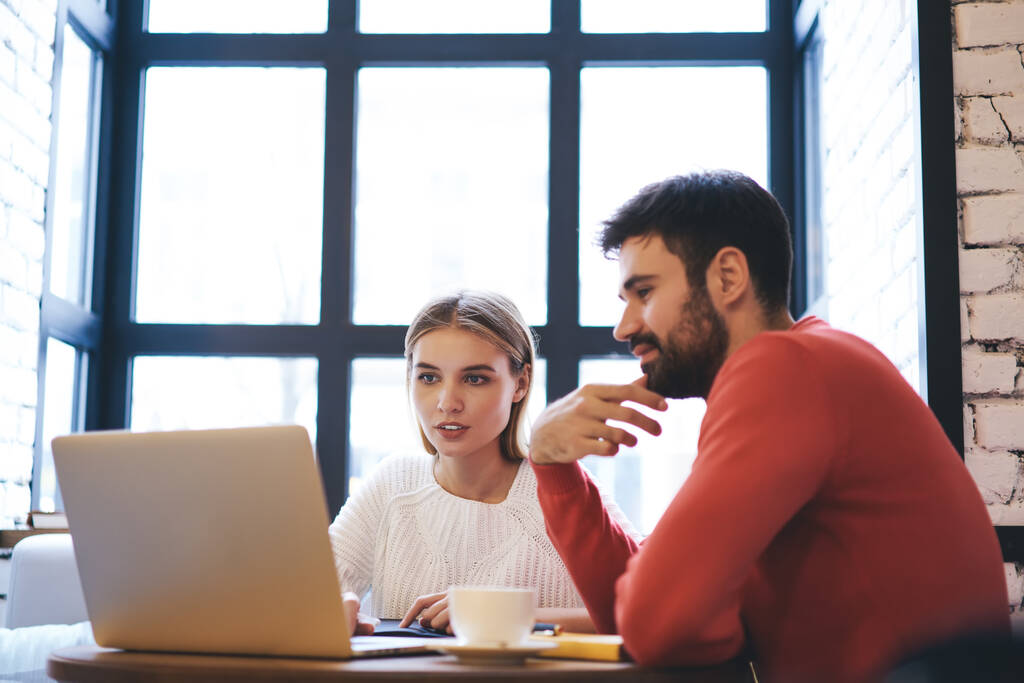 Moderne man vertelt te kalmeren vrouwelijke freelancer met laptop terwijl het kijken naar het scherm en zitten aan tafel met kopje in cafe overdag - Foto, afbeelding