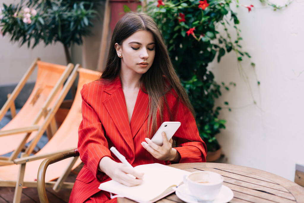 ストリートカフェのテーブルに座って携帯電話を拾い読みしながらプロジェクトに取り組んでスタイリッシュなスーツの深刻な若いビジネスマンの女性 - 写真・画像
