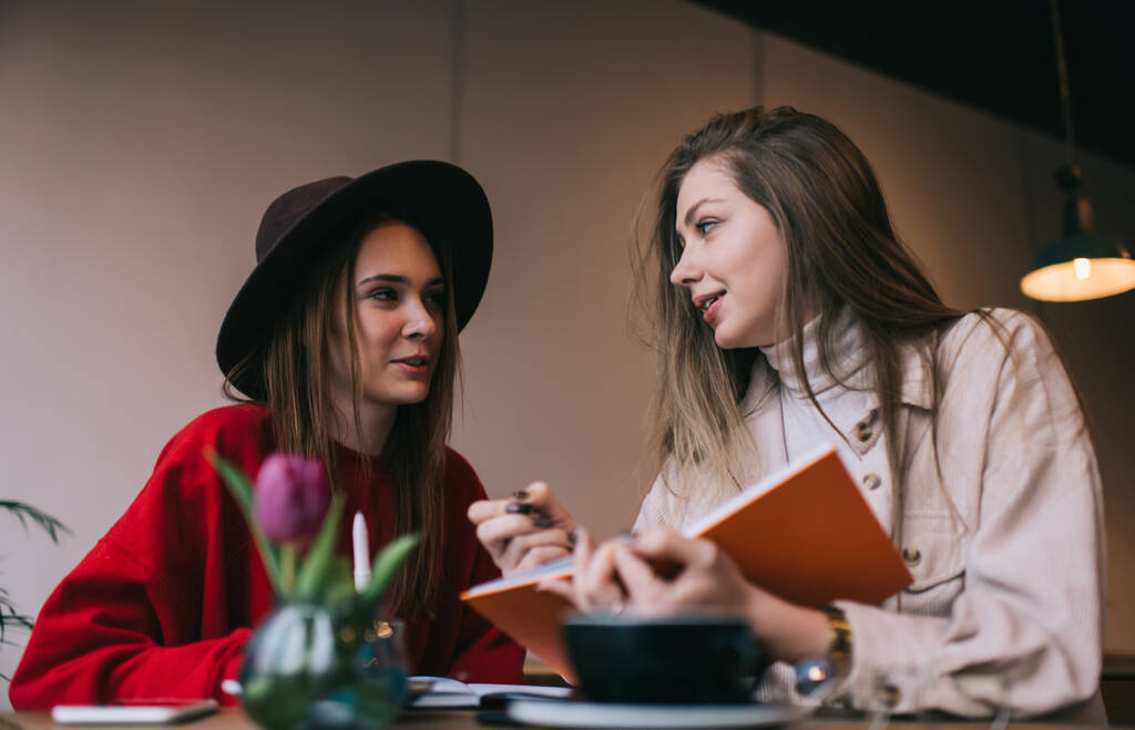 Задумчивые девушки в стильных нарядах, сидящие за деревянным столом и обсуждающие детали проекта во время дневного отдыха в уютном кафе - Фото, изображение