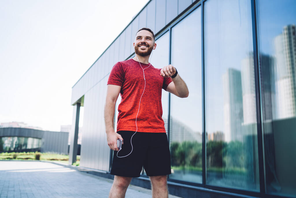 Nízký úhel veselého mužského atleta ve sportovním oblečení se sluchátky a chytré hodinky stojící na ulici při přípravě na běh - Fotografie, Obrázek