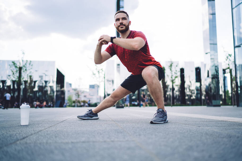 Full body of concentrated bearded sportsman in activewear and sneakers standing on paved street near plastic bottle of water and stretching legs before workout - Zdjęcie, obraz