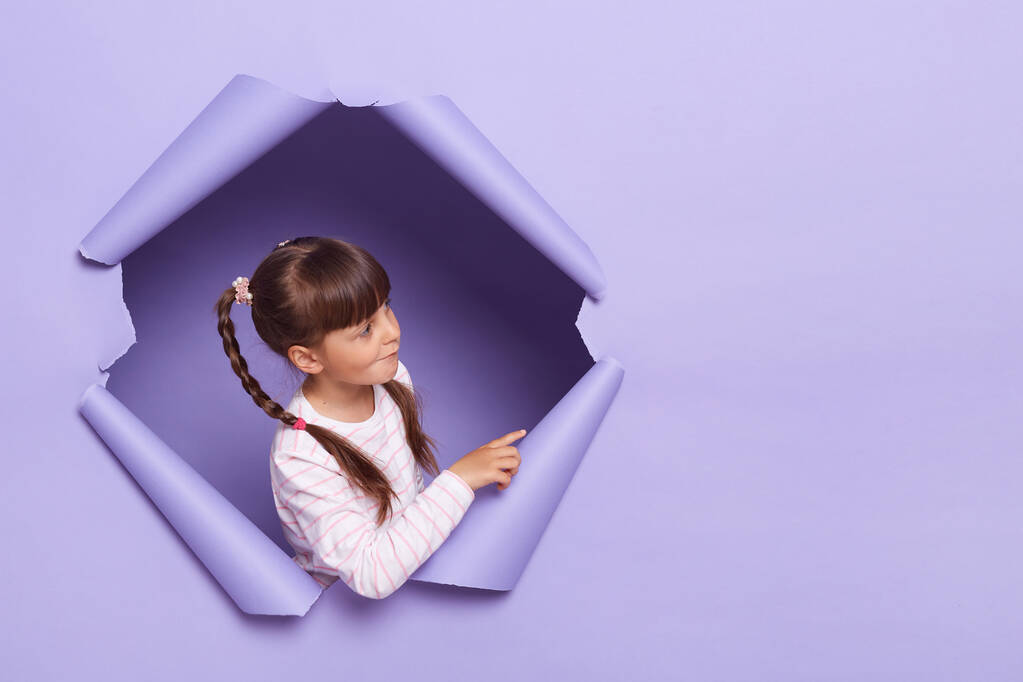Obraz ciemnowłosej dziewczynki ubranej w koszulkę w paski, pozującej w rozdarty fioletowy otwór papierowy, stojącej z boku na przestrzeń reklamową lub promocyjną. - Zdjęcie, obraz