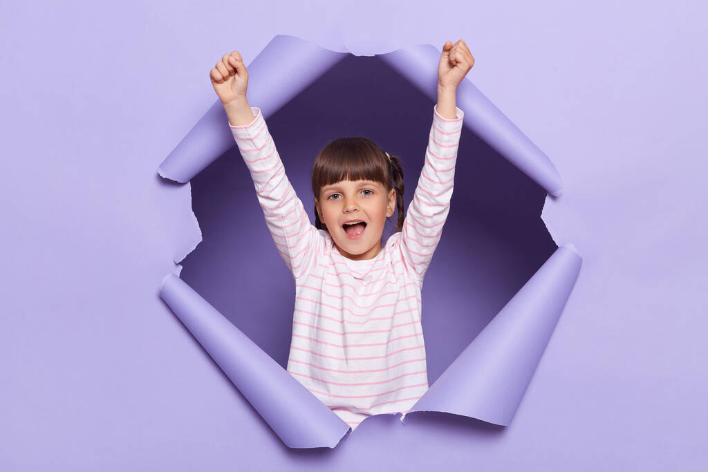 Horizontal shot of cute happy excited little girl wearing striped shirt standing with raised arms, screaming hurray, celebrating victory, posing in torn purple paper hole. - Photo, Image