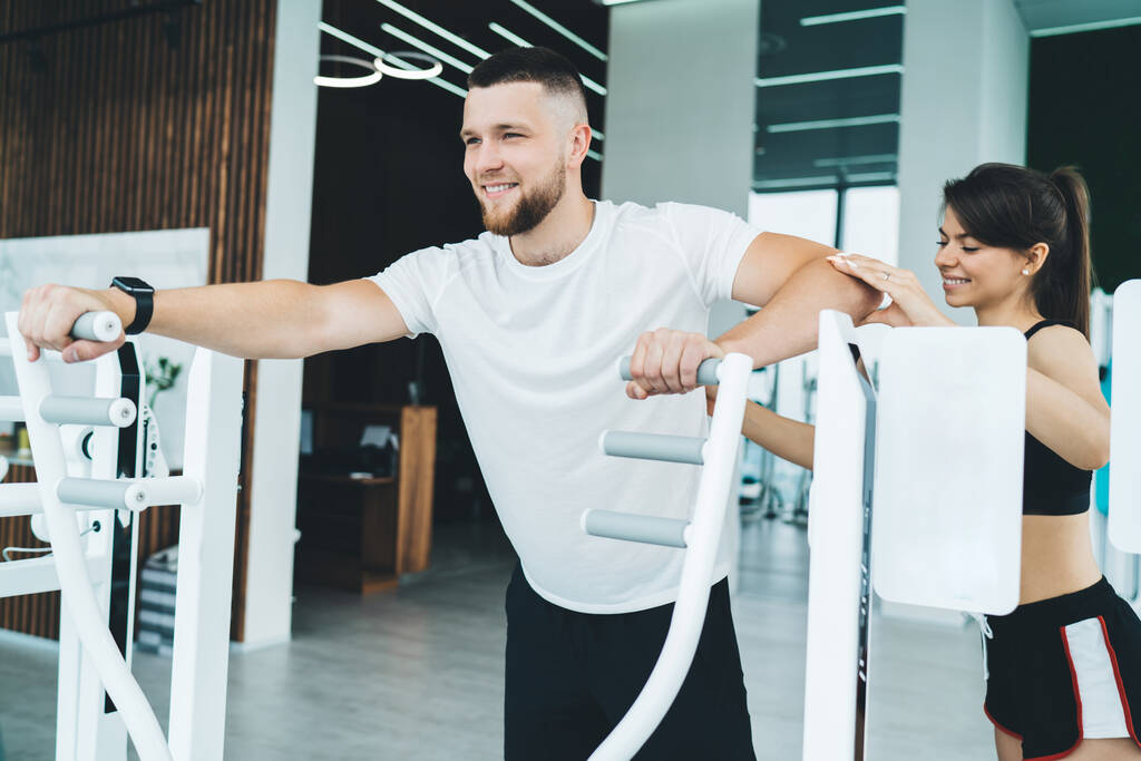 Positive Sportlerin in aktiver Kleidung, die Arme eines bärtigen Sportlers stützt, der während des Fitnesstrainings an modernen Sportgeräten trainiert - Foto, Bild